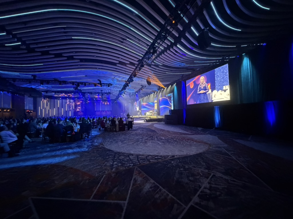 Wide view of the ballroom under blue wash with IMAG screens and banquet rounds